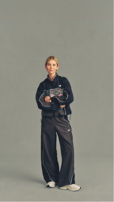 Full-body studio image of a person in a black New Balance tracksuit holding a patterned notebook against a neutral gray background.
