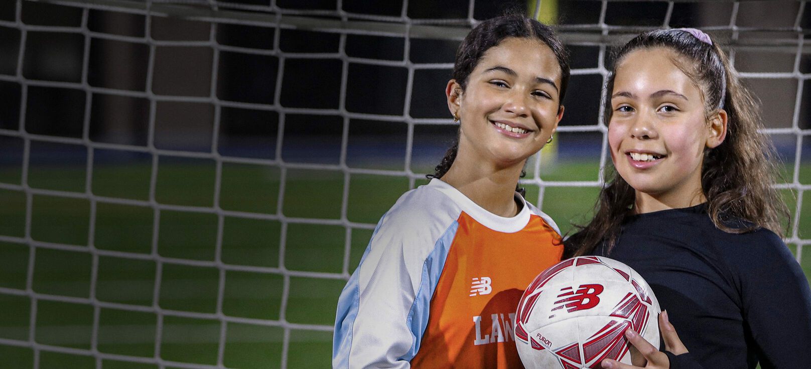 Two smiling teen girls posing with a soccer ball in front of a goal.