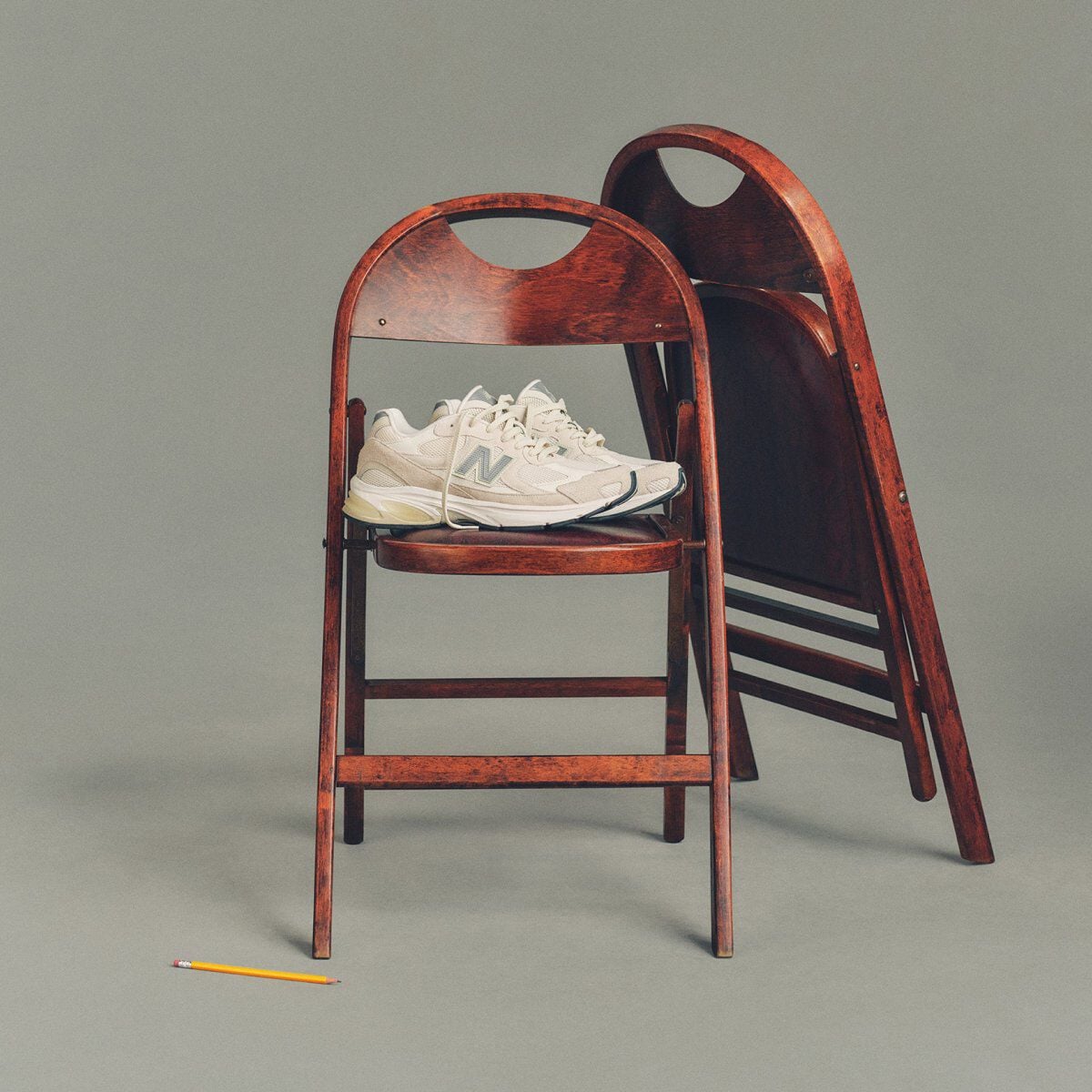 Studio image of a pair of cream-colored New Balance sneakers placed on a dark wooden folding chair, with a second matching chair folded and leaned behind it.