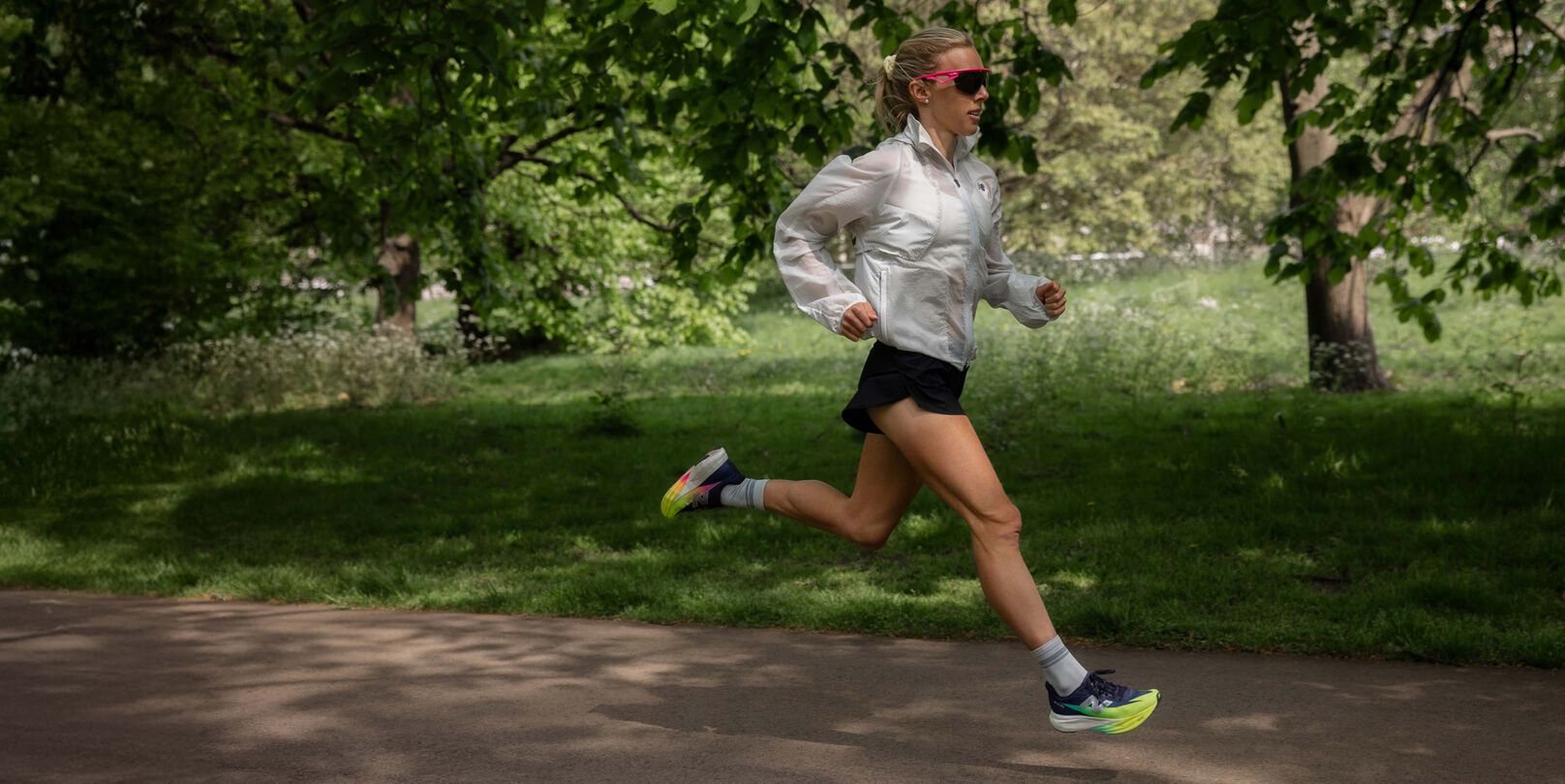 Emily Sisson running outdoors on a paved path in a park, wearing a white jacket, black shorts, and brightly colored running shoes. 