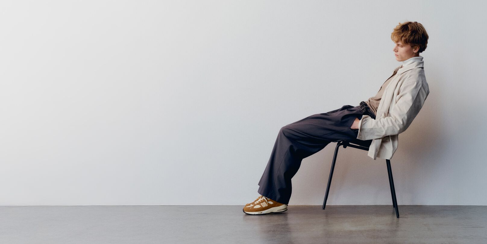 A man wearing a pair of New Balance Made in UK sneakers sitting on a chair leaning back in a room with a concrete floor and white walls. 