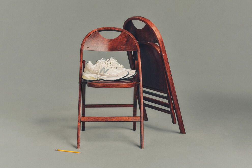 Studio image of a pair of cream-colored New Balance sneakers placed on a dark wooden folding chair, with a second matching chair folded and leaned behind it.